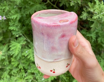 Könnte beinhalten: Eine rosa-weiße Keramiktasse mit einem Kirschendesign. Die Tasse hat ein kleines Loch oben für einen Strohhalm.