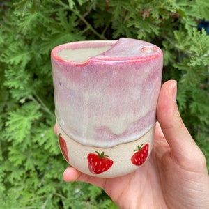 Puede incluir: Una taza de cerámica rosa y blanca con un diseño de fresa. La taza tiene una forma única con un pequeño labio en un lado.