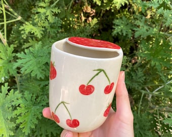 Op de afbeelding: Een witte keramische mok met een rood deksel en een kersenpatroon. De mok is versierd met rode kersen op een witte achtergrond.