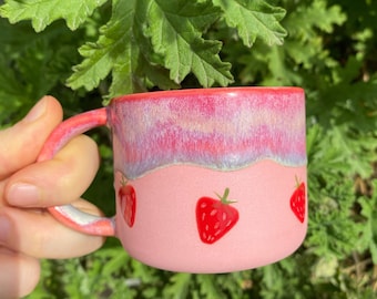 Könnte beinhalten: Eine rosa Keramiktasse mit weißem Rand und drei roten Erdbeermotiven. Die Tasse hat eine hellrosa Glasur mit einem dezenten irisierenden Finish.