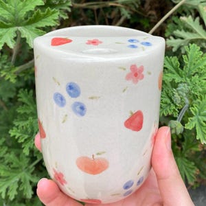 May include: A cylindrical ceramic cup with a cream-colored base, adorned with hand-painted red strawberries, blue blueberries, pink flowers, and orange apples. The cup is held in a hand against a backdrop of green foliage.