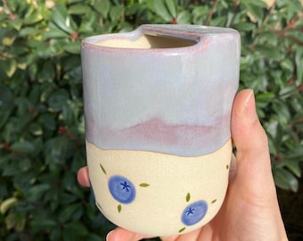 Könnte beinhalten: Eine handgefertigte Keramik-Tasse mit hellblau und lila glasierter Oberseite und cremefarbenem Boden. Der Boden ist mit blauen Blaubeer-Illustrationen und grünen Blättern verziert. Die Tasse hat ein einzigartiges Griffdesign.