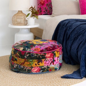 Könnte beinhalten: Ein runder, mehrfarbiger Patchwork-Hocker mit rosa Samtbesatz steht auf einem gewebten Teppich vor einem Bett mit einer dunkelblauen Tagesdecke und rosa und beigen Kissen.