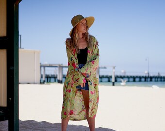 Könnte beinhalten: Eine Frau in einem grünen Blumen-Kimono mit einem breitkrempigen Hut steht an einem Sandstrand.