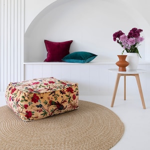 Könnte beinhalten: Ein floral gemusterter Ottoman mit einem runden Jute-Teppich davor. Zwei Kissen liegen auf einem weißen Regal hinter dem Ottoman, eines ist türkis und das andere burgunderrot. Ein kleiner Holztisch mit einer weißen Platte trägt eine Vase mit lila Blumen.