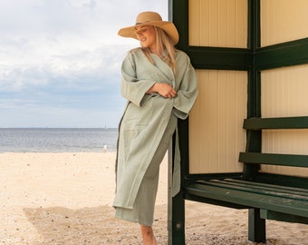 Op de afbeelding: Een vrouw in een lichtgroene badjas en een strohoed staat in een strandhut. De hut heeft een houten bank en een groen dak.