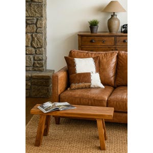 May include: A living room scene featuring a brown leather sofa with decorative pillows. A small wooden bench holds an open book. A stone fireplace and wooden dresser with a lamp and plant complete the cozy interior.