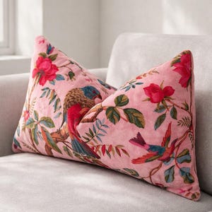 May include: Pink cushion with a floral and bird pattern. The cushion features a repeating design of red roses, green leaves, and colourful birds. The cushion is rectangular and rests on a light grey sofa.