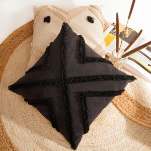 May include: Two decorative throw pillows with a geometric pattern. The pillow on the bottom is black with black fringe and the pillow on top is beige with white fringe.