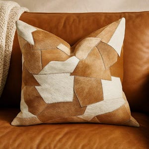 May include: A decorative square pillow with a patchwork design. The pillow features various shapes of tan, brown, and cream-colored leather pieces sewn together. The pillow is resting on a brown leather couch.