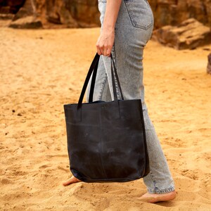 May include: A person carrying a black leather tote bag with a wide strap. The bag has a simple design and is made of a durable material.