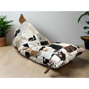 May include: A patchwork beanbag chair in a triangular shape. The chair is made of various cowhide patches in shades of brown, black, and white. A brown leather strap is attached to the side. The beanbag is on a light wood floor.
