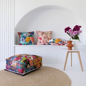 May include: A colorful patchwork floor cushion with a floral print sits on a beige woven rug in front of a white wall with a niche. Three floral patterned throw pillows are displayed on a shelf in the niche. A small wooden table with a white top holds a vase of purple flowers.