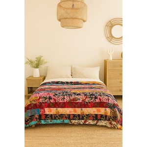 May include: A vibrant patchwork quilt featuring floral designs and horizontal stripes in pink, red, blue, and black. The quilt is placed on a bed with white pillows, and the room has a neutral colour scheme.
