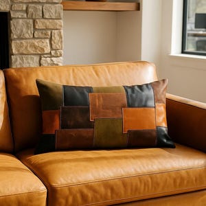 Peut inclure: Un coussin rectangulaire décoratif avec un motif patchwork dans les tons de marron, noir et vert olive repose sur un canapé en cuir beige. Le motif du coussin présente des pièces de cuir rectangulaires. Le canapé est dans un salon bien éclairé.
