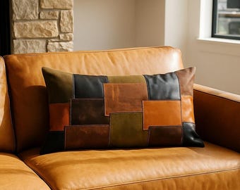 May include: A rectangular cushion with a patchwork design in shades of brown, black, and olive green rests on a tan leather sofa. The cushion's design features rectangular leather patches. The sofa is in a well-lit living room.