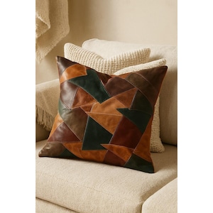 Peut inclure: Un coussin décoratif avec un motif patchwork. Le coussin présente des formes géométriques dans des tons de marron, de beige et de vert. Le coussin est placé sur un canapé beige, avec d'autres coussins en arrière-plan.