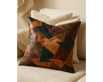 May include: A decorative cushion with a patchwork design. The cushion features geometric shapes in shades of brown, tan, and green. The cushion is placed on a beige sofa, with other cushions in the background.