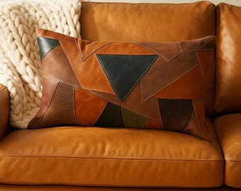 May include: A rectangular cushion with a patchwork design in shades of brown, tan, and black. The cushion is placed on a brown leather sofa, with a cream-coloured knitted throw blanket visible in the background.