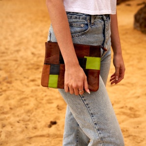 Puede incluir: Un embrague de cuero marrón con parches de color verde, negro y marrón. El embrague lo lleva una persona que lleva pantalones vaqueros azules.