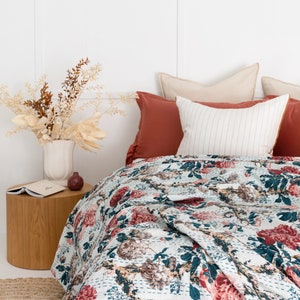 May include: A white bed with a floral patterned quilt and a red and beige pillow. The bed is against a white wall with a vase of dried flowers on a wooden side table.