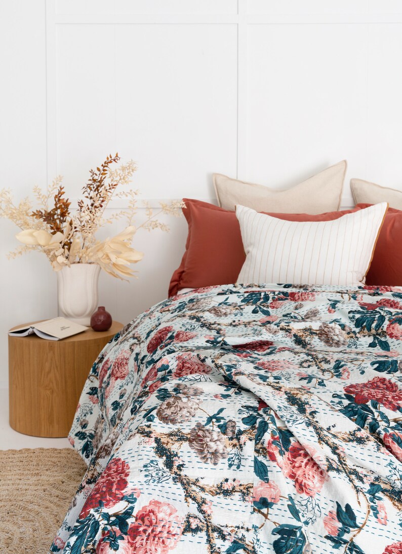 May include: A white bed with a floral patterned quilt and a red and beige pillow. The bed is against a white wall with a vase of dried flowers on a wooden side table.