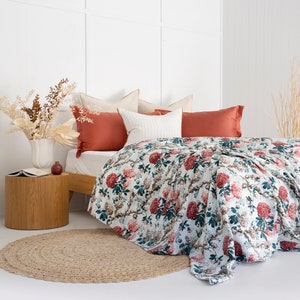 May include: A white bed with a floral patterned quilt and red and white pillows. The bed is in a white room with a wooden side table and a jute rug.