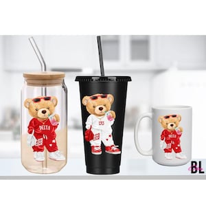 Puede incluir: Tres art&iacute;culos de cristaler&iacute;a con un dise&ntilde;o de oso de peluche. El oso lleva gafas de sol y un ch&aacute;ndal rojo con "DELTA" impreso. Los art&iacute;culos incluyen un vaso con tapa de bamb&uacute;, un vaso negro y una taza blanca.