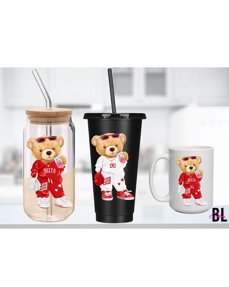 Puede incluir: Tres art&iacute;culos de cristaler&iacute;a con un dise&ntilde;o de oso de peluche. El oso lleva gafas de sol y un ch&aacute;ndal rojo con "DELTA" impreso. Los art&iacute;culos incluyen un vaso con tapa de bamb&uacute;, un vaso negro y una taza blanca.