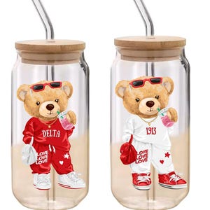 Puede incluir: Dos vasos de vidrio transparente con tapas de madera y pajitas de vidrio. Cada vaso presenta un oso de peluche de dibujos animados con gafas de sol y un ch&aacute;ndal. Un oso viste de rojo, el otro de blanco. El texto incluye "DELTA", "1913" y "LOVE LOVE LOVE".