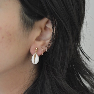 Peut inclure: Une paire de boucles d'oreilles avec un petit charme en forme de coquillage blanc suspendu à un clou en or. Les boucles d'oreilles sont portées à l'oreille gauche de la personne.