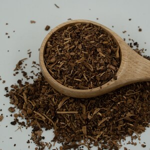 May include: A wooden spoon filled with brown, dried, and shredded herbs. The spoon is resting on a white surface with a small amount of the herbs scattered around it.