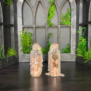 May include: Two clear glass bottles filled with pink Himalayan salt and topped with a wax seal. Each bottle has a silver key charm hanging from the top. The bottles are sitting on a dark surface in front of a window with a gothic design.