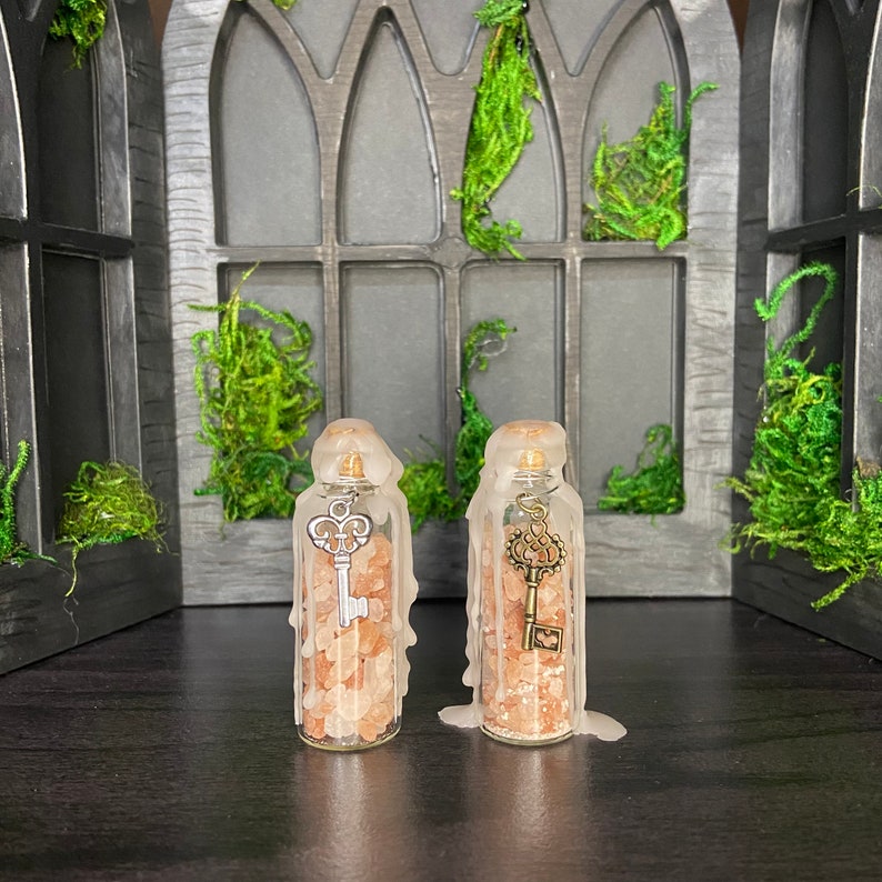 May include: Two clear glass bottles filled with pink Himalayan salt and topped with a wax seal. Each bottle has a silver key charm hanging from the top. The bottles are sitting on a dark surface in front of a window with a gothic design.