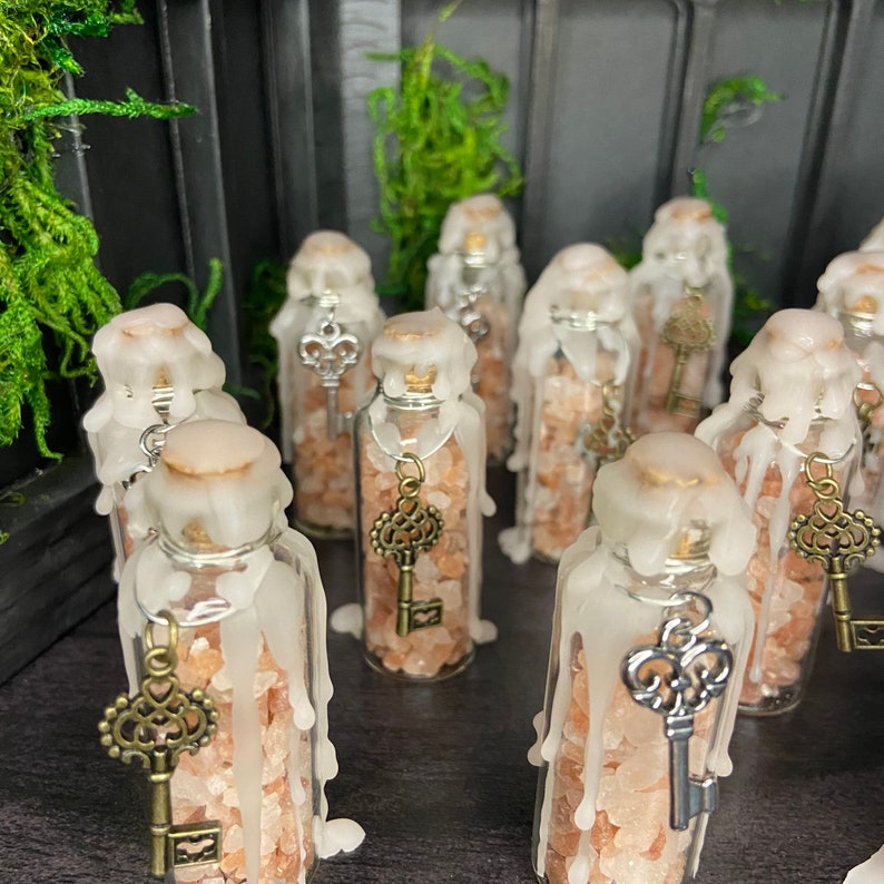 May include: A collection of small glass bottles filled with pink Himalayan salt and topped with white wax. Each bottle has a silver key charm hanging from it.