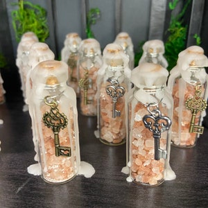 May include: A collection of small glass bottles filled with pink Himalayan salt and topped with a white wax drip. Each bottle has a silver key charm hanging from the top.