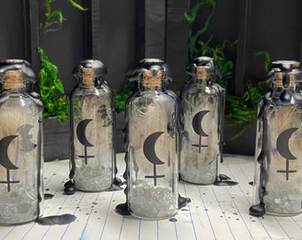 May include: Five small glass bottles with cork stoppers are filled with glitter and a white feather. Each bottle is sealed with black wax and has a black and white symbol of a crescent moon and a cross on the front.