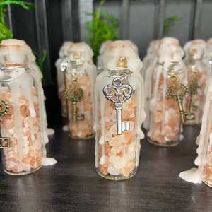 May include: A collection of small glass bottles filled with pink Himalayan salt and topped with a white wax candle. Each bottle has a silver key charm hanging from the top.