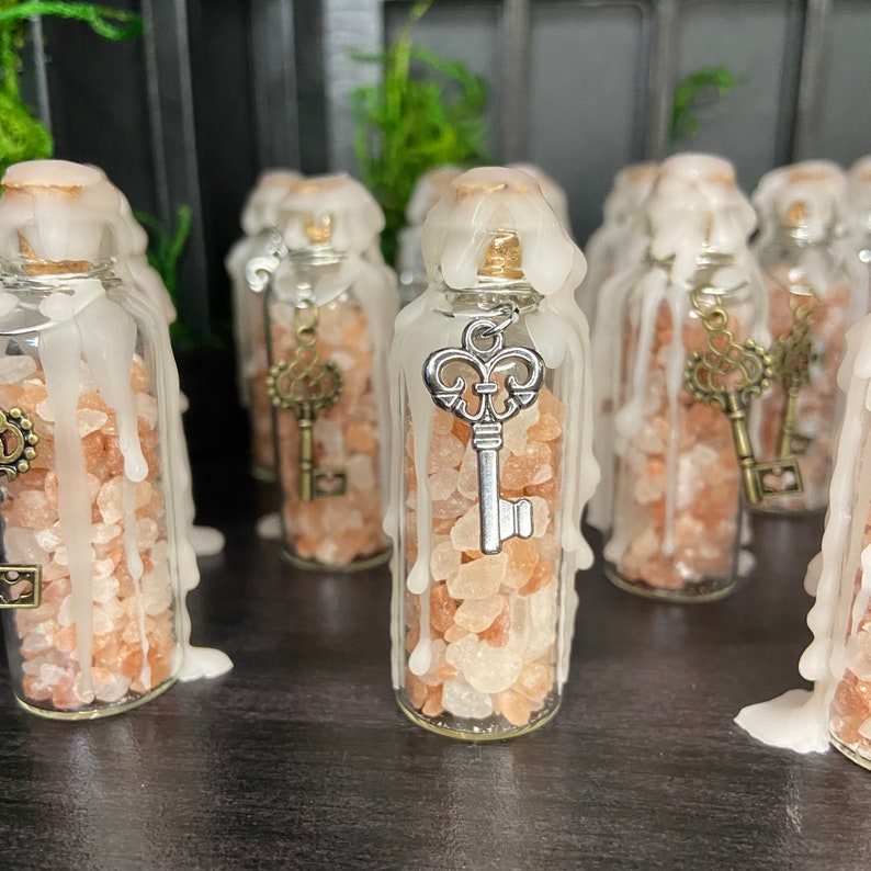 May include: A collection of small glass bottles filled with pink Himalayan salt and topped with a white wax candle. Each bottle has a silver key charm hanging from the top.