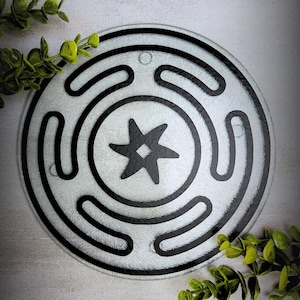 May include: A circular glass cutting board featuring a textured surface with a black printed design. The pattern consists of a central six-pointed star enclosed within concentric circular lines and curved segments, creating a geometric motif.