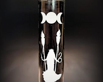 May include: A tall, cylindrical black glass candle featuring a white graphic print. The design includes a triple moon symbol at the top, positioned above a central figure wearing a crown and holding two tall, flaming torches on either side.