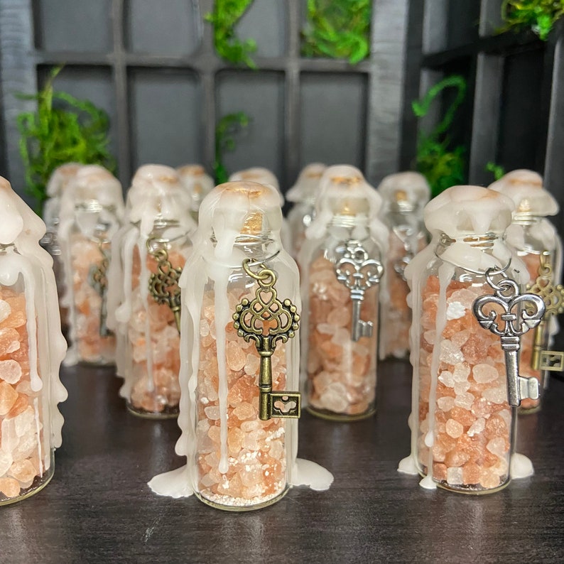 May include: A collection of small glass bottles filled with pink Himalayan salt and topped with a white wax seal. Each bottle has a small key charm hanging from the top.