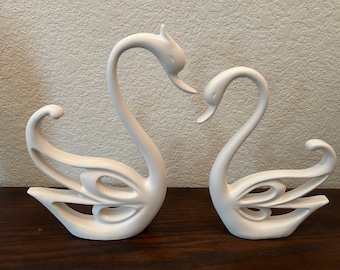 May include: A pair of white ceramic swan figurines with intricate designs. The swans are facing each other with their necks curved.