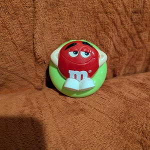May include: A red M&M's candy character with a smiling face, white shoes, and a green base. The candy character has the letter "m" on its front. The image is set against a brown, textured background.