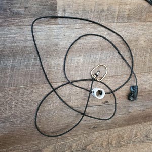 May include: A black rope with a gold-colored ring, a white plastic piece, and a black plastic clip. The rope is lying on a wooden surface.