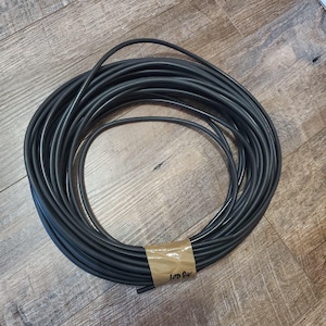 May include: A coiled black cable, likely for electrical or data transmission, is shown on a wooden surface. The cable is neatly wound and secured with a brown paper band. The band has the text "100 ft" printed on it.