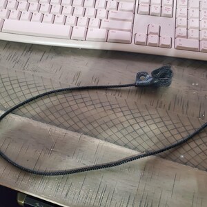 May include: A black leather pull tab with a braided black cord, likely for a zipper or similar application. The pull tab is attached to the cord with a small metal rivet. The background includes a pink keyboard and a wooden surface.