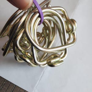 May include: A tangled bundle of shiny, gold-colored metal loops, held together by a purple rubber band. The loops are curved and appear to be made of a sturdy material. The background is a white surface.