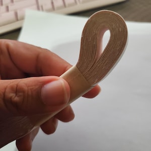 May include: A roll of beige paper tape, looped to show the layers. The tape is held in a hand, with a white surface and a keyboard in the background.