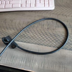 May include: A black, flexible cable with a leather loop at one end. The cable is curved and lies on a wooden surface. A pink keyboard is visible in the background.